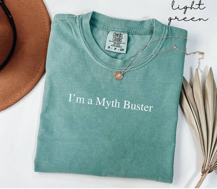 Folded light green t-shirt with 'I'm a Myth Buster' text, next to a brown hat and dried leaves.