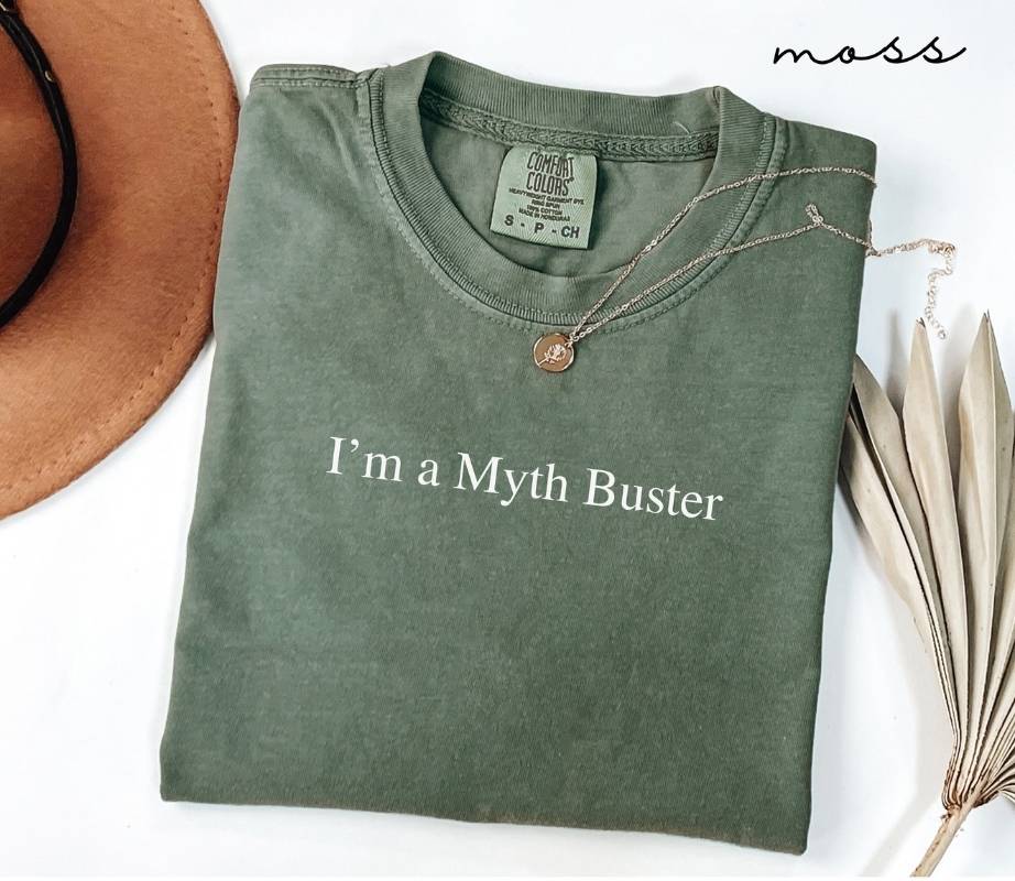 Folded green (moss) t-shirt with 'I'm a Myth Buster' text, placed on a white surface with a hat and dried leaves.