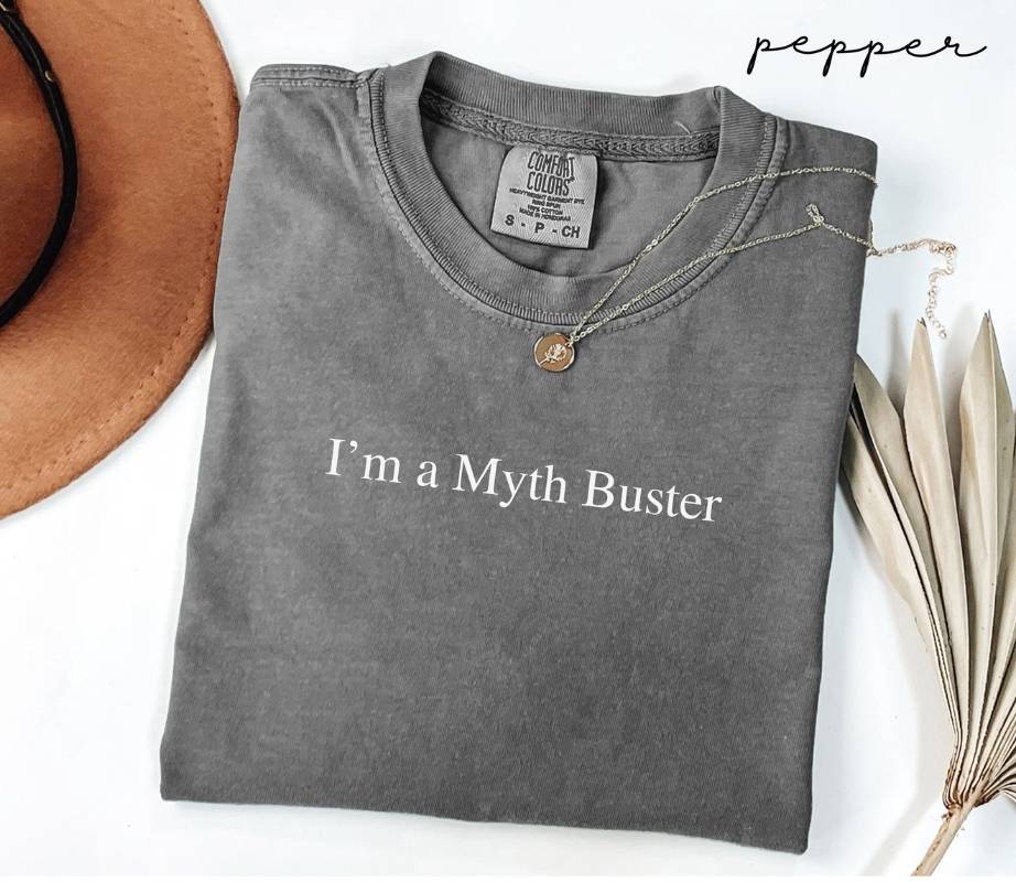 Folded gray (pepper) t-shirt with 'I'm a Myth Buster' text, next to a hat and dried leaves on a white background.