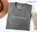 Folded gray (pepper) t-shirt with 'I'm a Myth Buster' text, next to a hat and dried leaves on a white background.