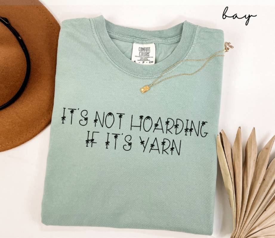 Light green (bay) t-shirt with text 'It's not hoarding if it's yarn' on a white background.