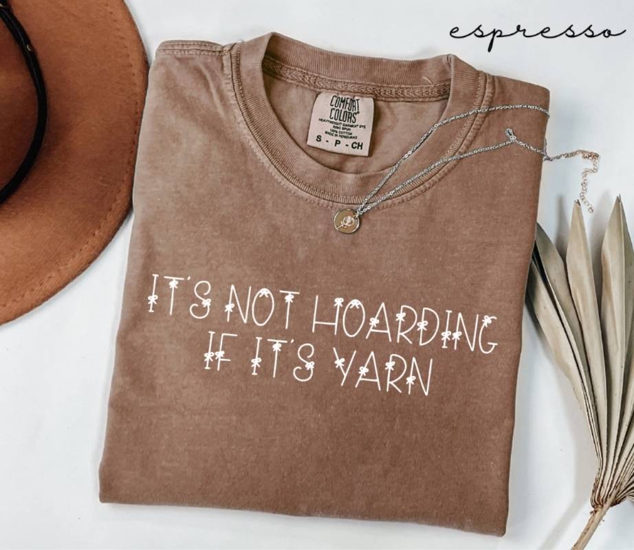 Folded brown (espresso) t-shirt with 'It's not hoarding if it's yarn' text, next to a hat and dried leaves on a white surface.