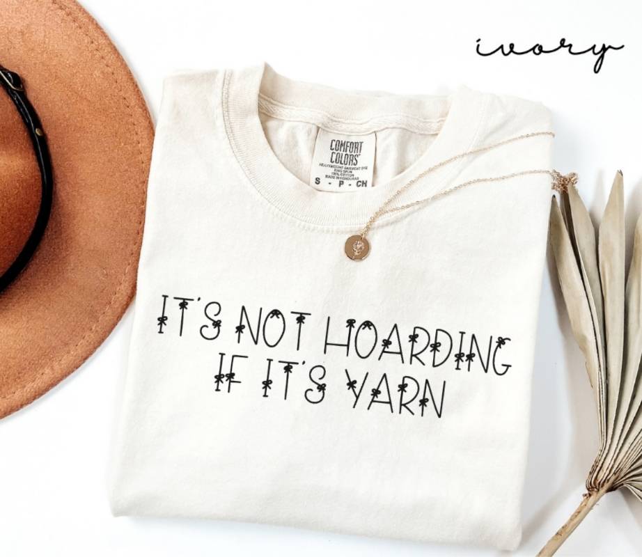White (ivory) t-shirt with text 'It's not hoarding if it's yarn' on a white background with a hat and dried leaves.