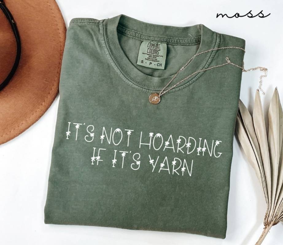 Folded green (moss) t-shirt with 'It's not hoarding if it's yarn' text, next to a brown hat and dried leaves on a white background.