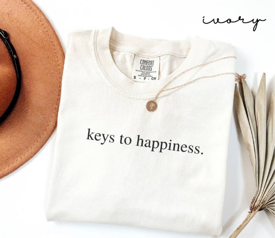 Folded white (ivory) t-shirt with 'keys to happiness.' text, next to a brown hat and dried plant on a white background.