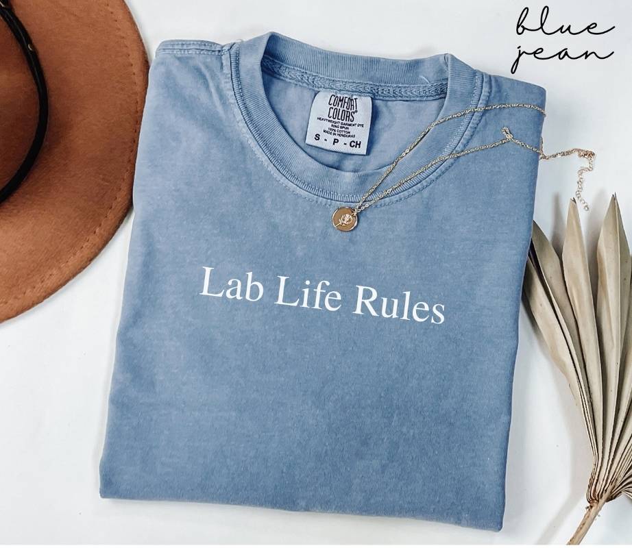 Folded blue jean t-shirt  with 'Lab Life Rules' text, necklace, and hat on a white background