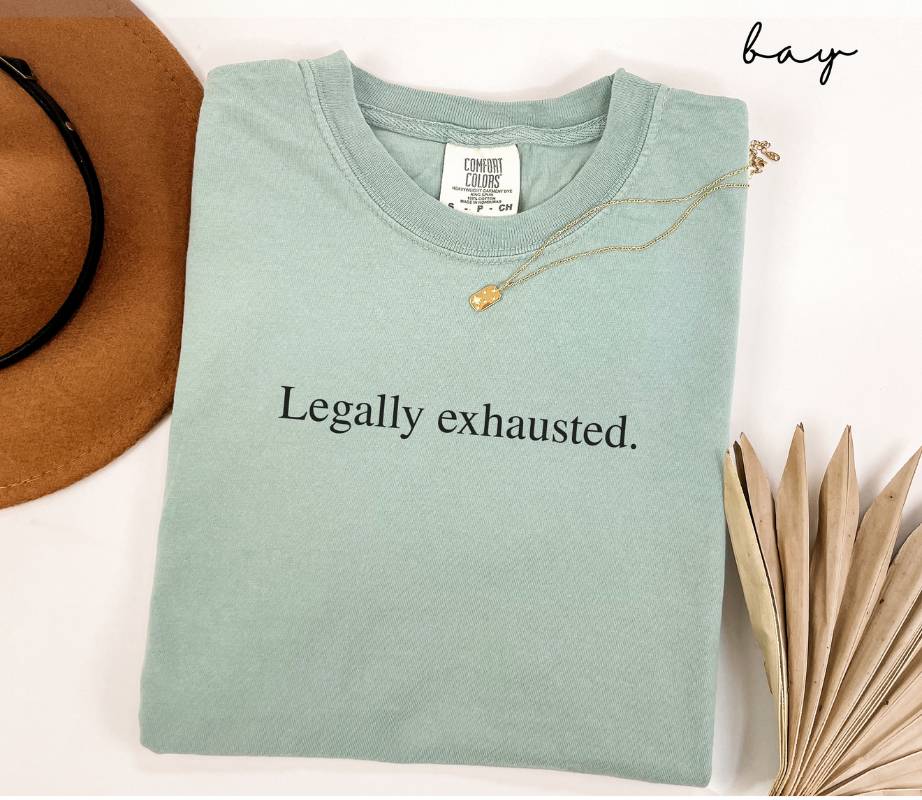 Folded light green (bay) t-shirt with 'Legally exhausted.' text, next to a hat and dried leaves on a white background.