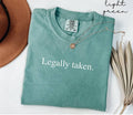 Folded light green t-shirt with 'Legally taken.' text, next to a brown hat and dried leaves.