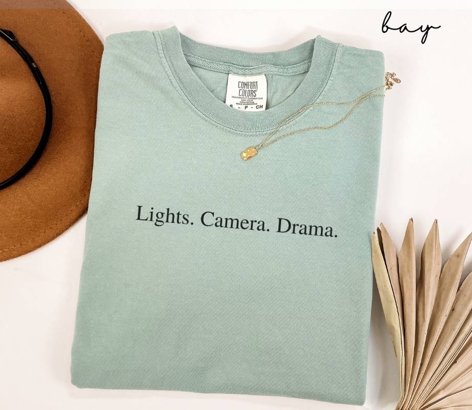 Folded light green (bay) t-shirt with 'Lights. Camera. Drama.' text, next to a brown hat and dried leaves on a white background.