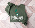Military green sweatshirt with the text 'Macau is calling, and I must go' on a wooden floor.