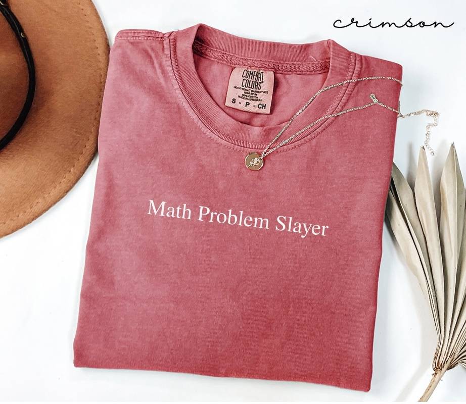 Folded pink (crimson) t-shirt with 'Math Problem Slayer' text, placed on a white surface with a hat and dried leaves.