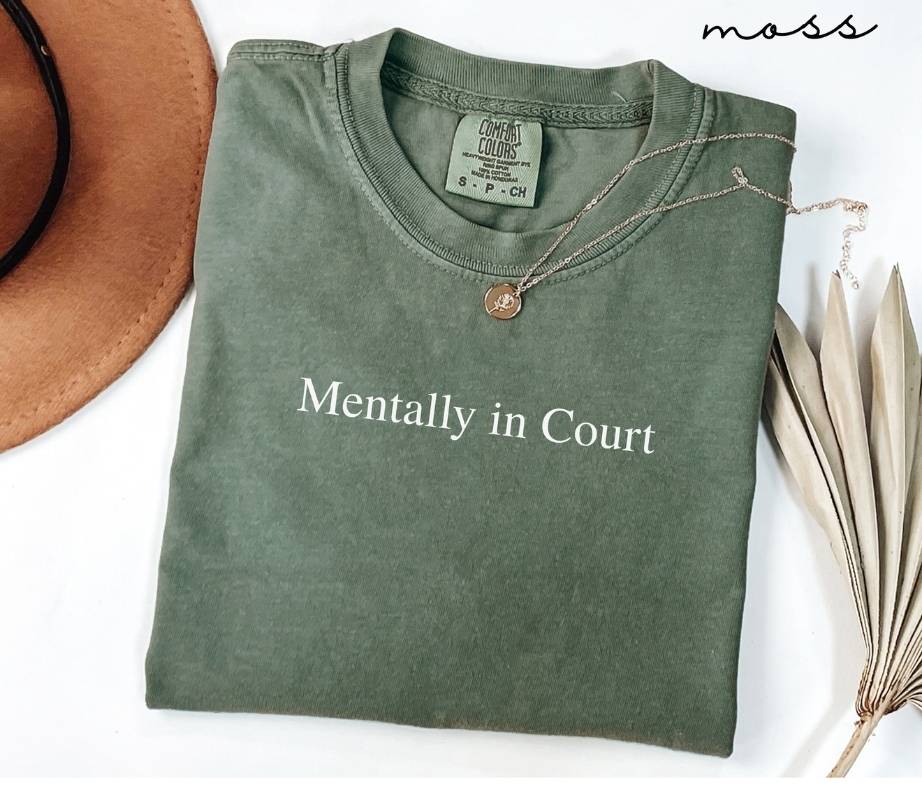 Folded green (moss) t-shirt with 'Mentally in Court' text, next to a brown hat and dried leaves on a white background.