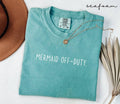 Folded Blue (seafoam) t-shirt with 'Mermaid Off-Duty' text, surrounded by a hat and dried leaves on a white background.
