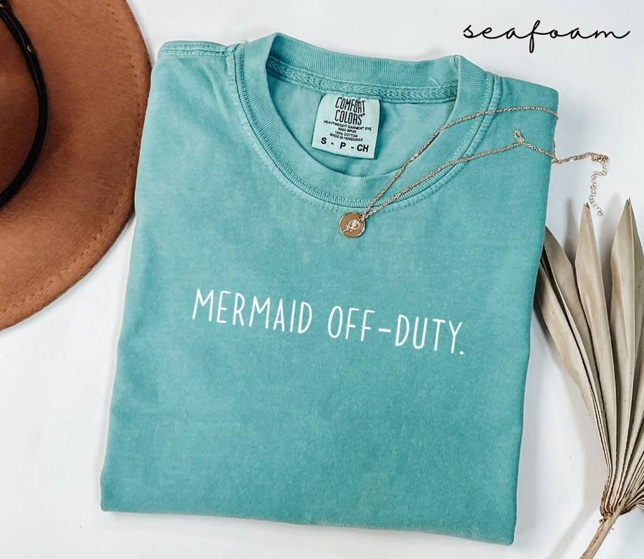 Folded Blue (seafoam) t-shirt with 'Mermaid Off-Duty' text, surrounded by a hat and dried leaves on a white background.