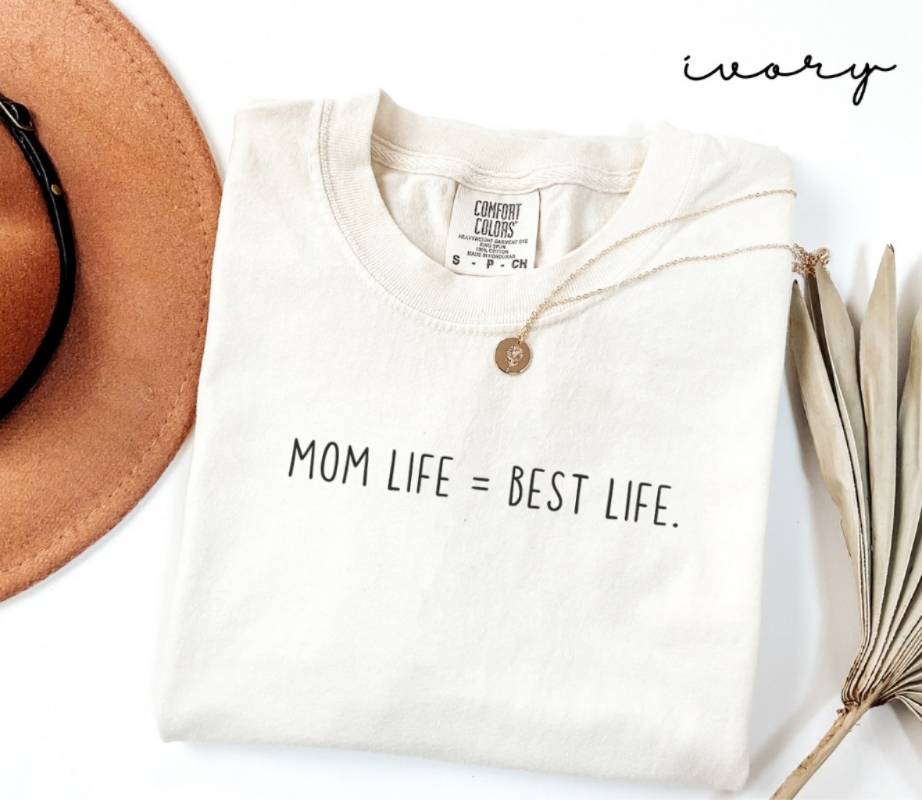 White t-shirt with the text 'Mom Life = Best Life', placed on a white surface alongside a hat and a dried plant.