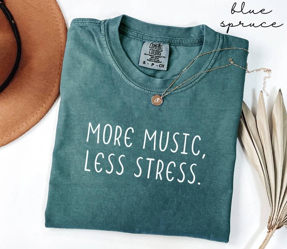 Folded teal (blue spruce) t-shirt with 'More Music, Less Stress' text on a white background with a hat and dried leaves.