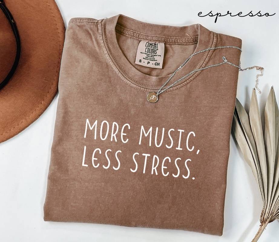 Folded brown (espresso) t-shirt with 'More Music, Less Stress' text on a white surface with a hat and dried leaves.