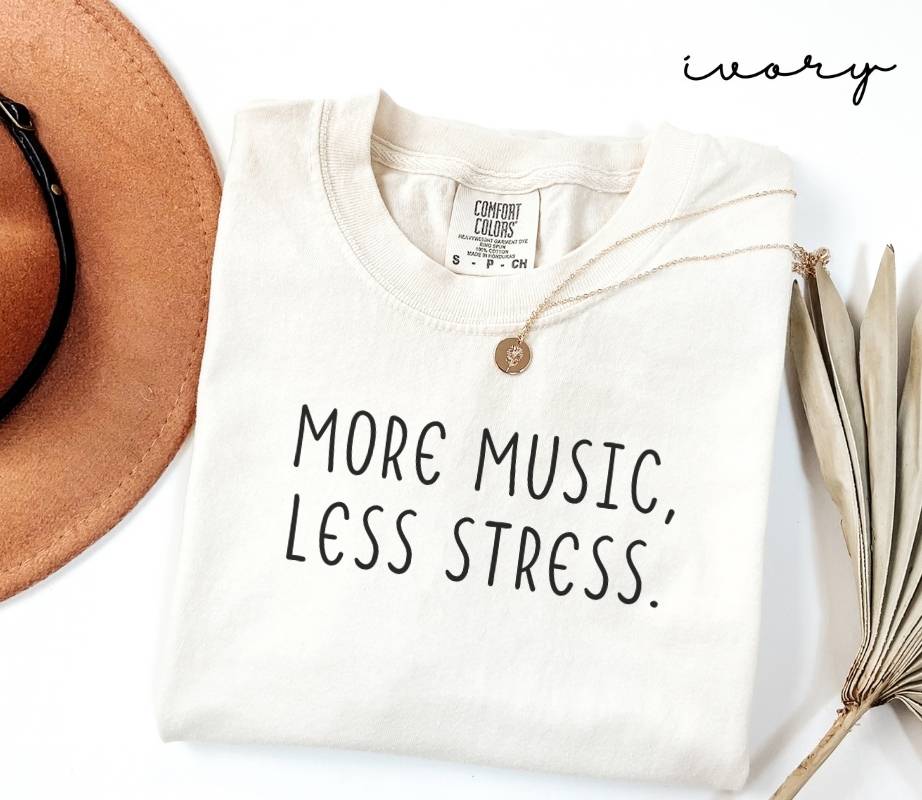 White (ivory) t-shirt with 'More Music, Less Stress' text, placed on a white surface with a hat and dried plant.