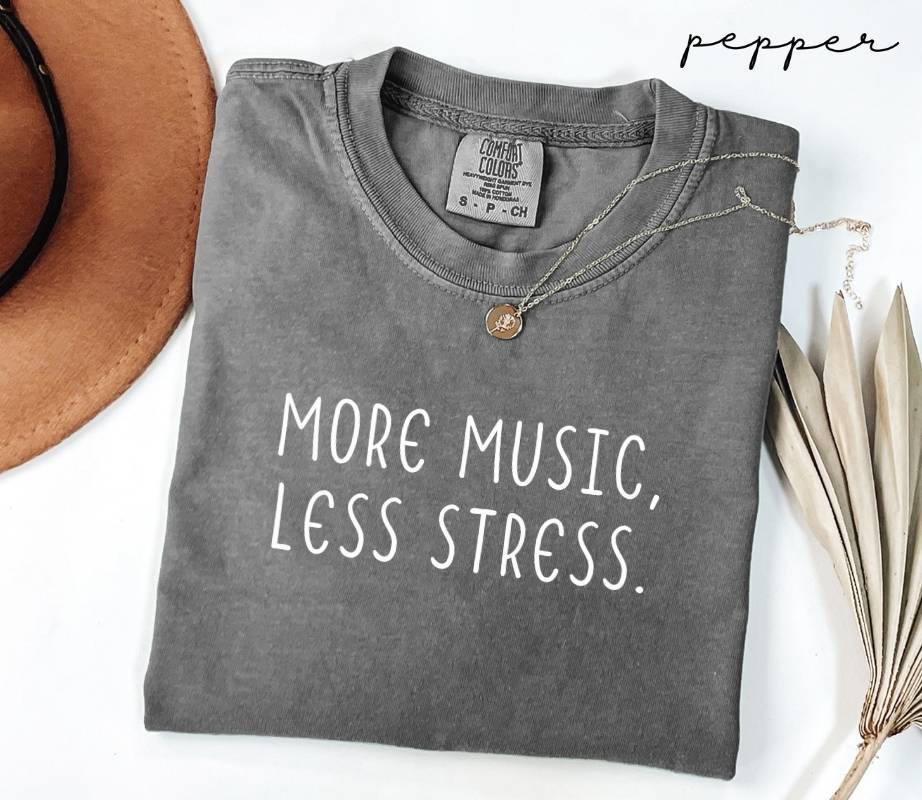 Folded gray (pepper) t-shirt with 'More Music, Less Stress' text on a white surface with a hat and dried leaves.