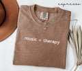 Folded brown (espresso) t-shirt with 'music = therapy' text, next to a hat and dried leaves on a white surface.