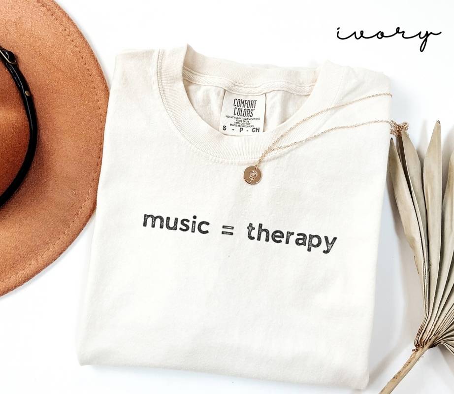 White (ivory) t-shirt with 'music = therapy' text, beige hat, and dried plant on a white background
