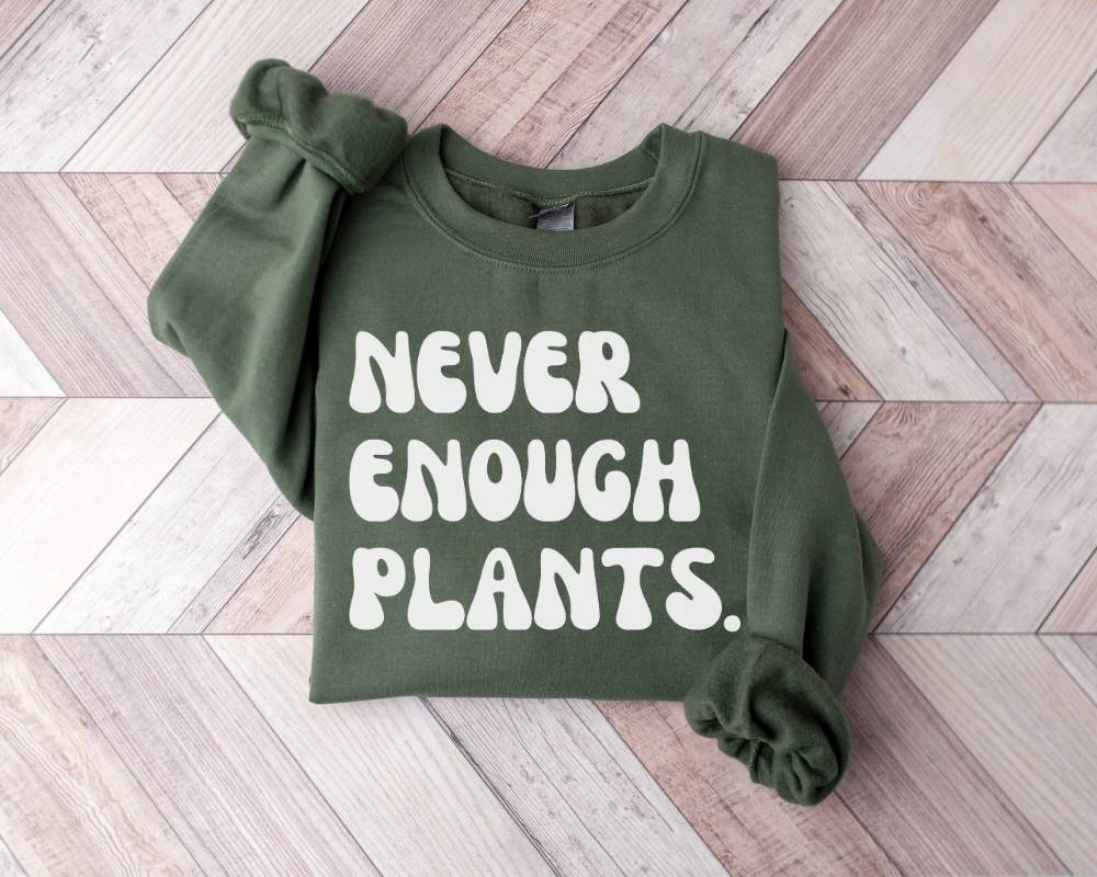 Military green sweatshirt with 'NEVER ENOUGH PLANTS' text on a wooden floor