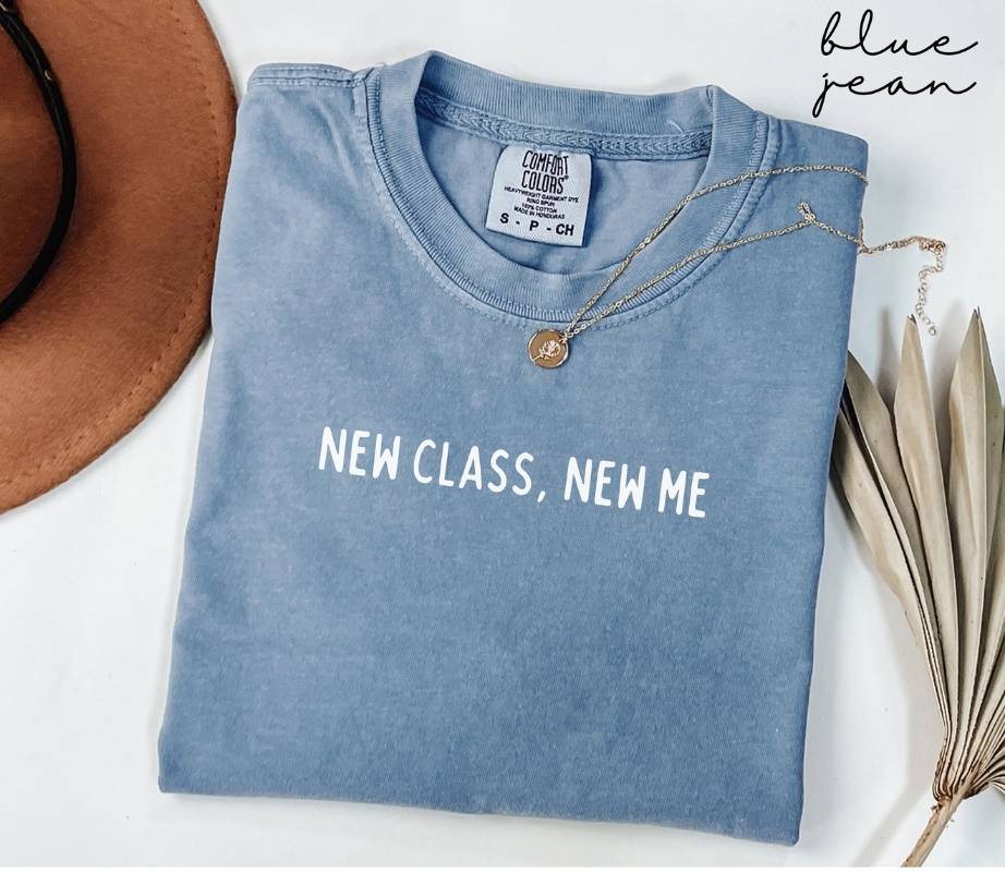 Folded blue jean t-shirt with 'NEW CLASS, NEW ME' text, necklace, and hat on a white background