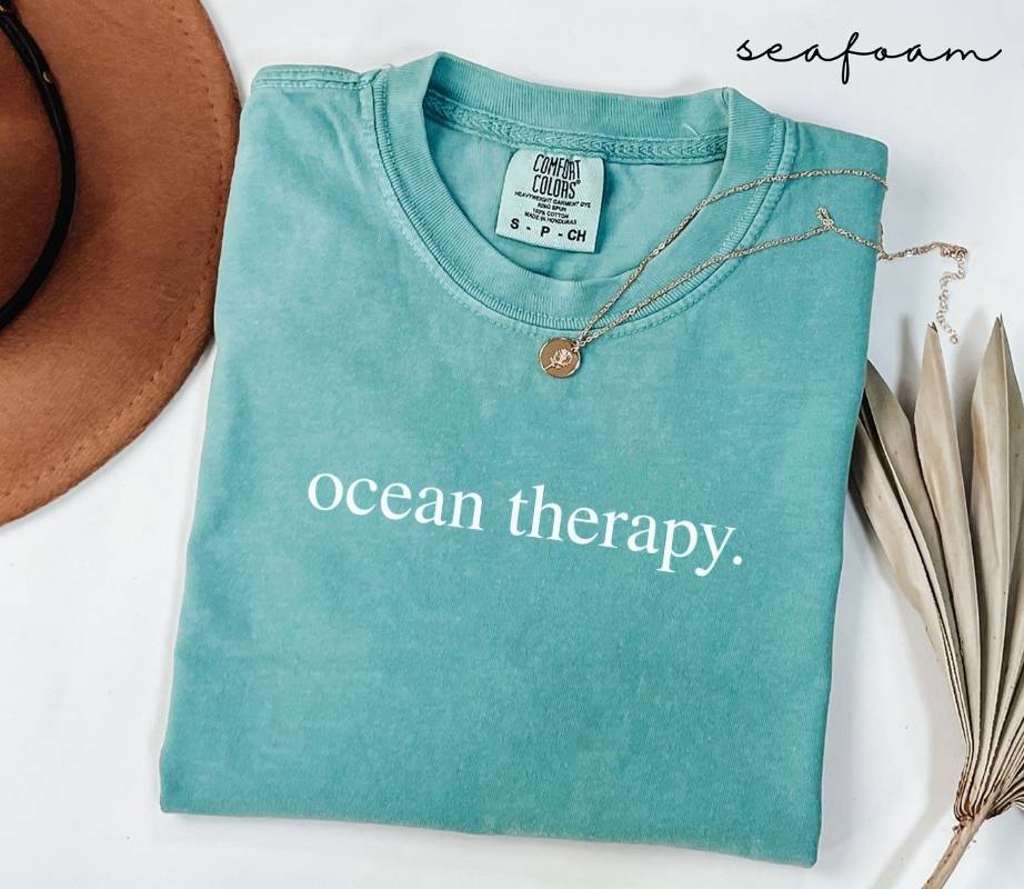 Folded teal (seafoam) t-shirt with 'ocean therapy.' text, surrounded by a hat and dried leaves on a white background.