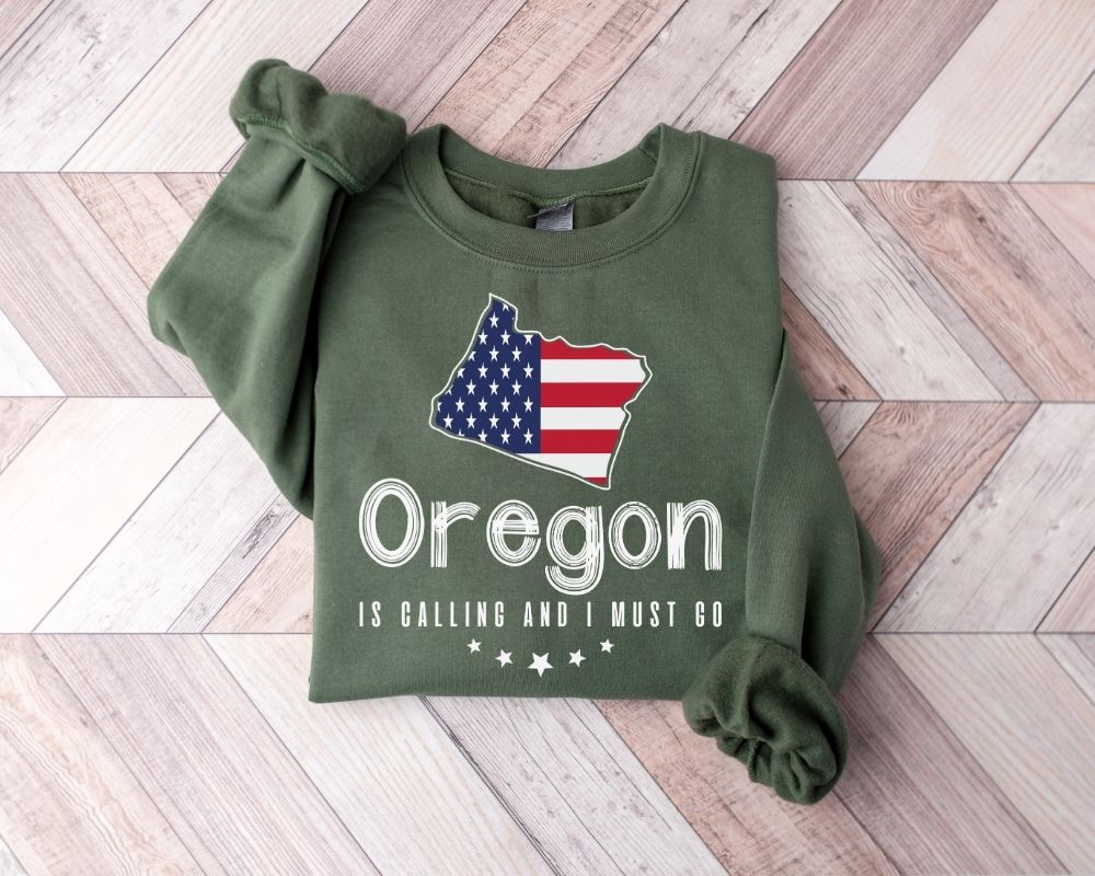 Military Green sweatshirt featuring the design 'Oregon is Calling and I Must Go' on a wooden floor.