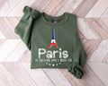 Military green sweatshirt featuring an Eiffel Tower design with the text 'Paris is Calling' placed on a wooden floor.