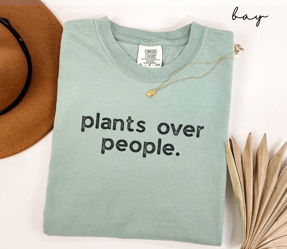 Folded light green (bay) t-shirt with 'plants over people.' text, next to a hat and dried leaves on a white background.