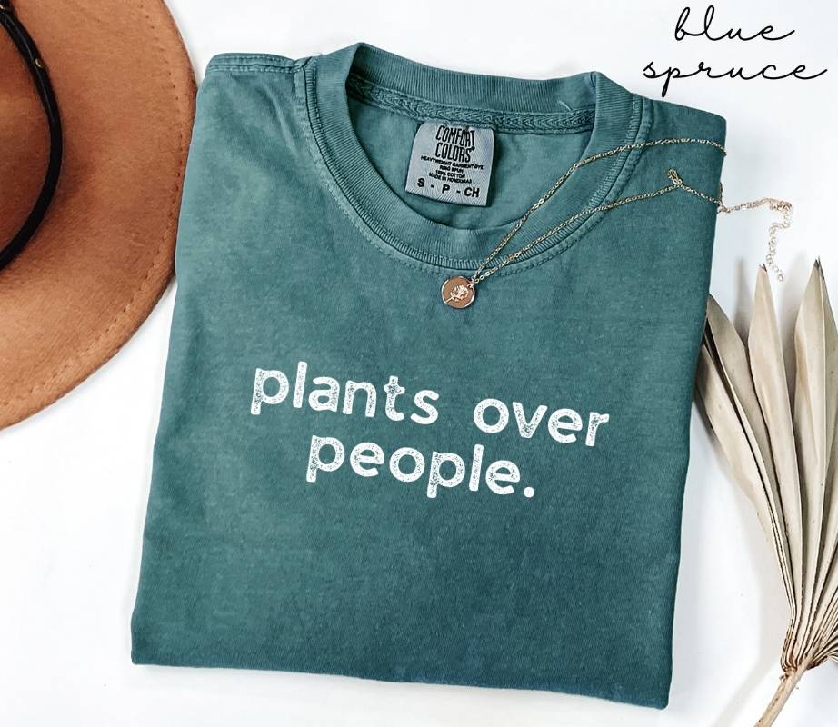 Folded teal (blue spruce) t-shirt with 'plants over people.' text, next to a brown hat and dried plant leaves on a white background.