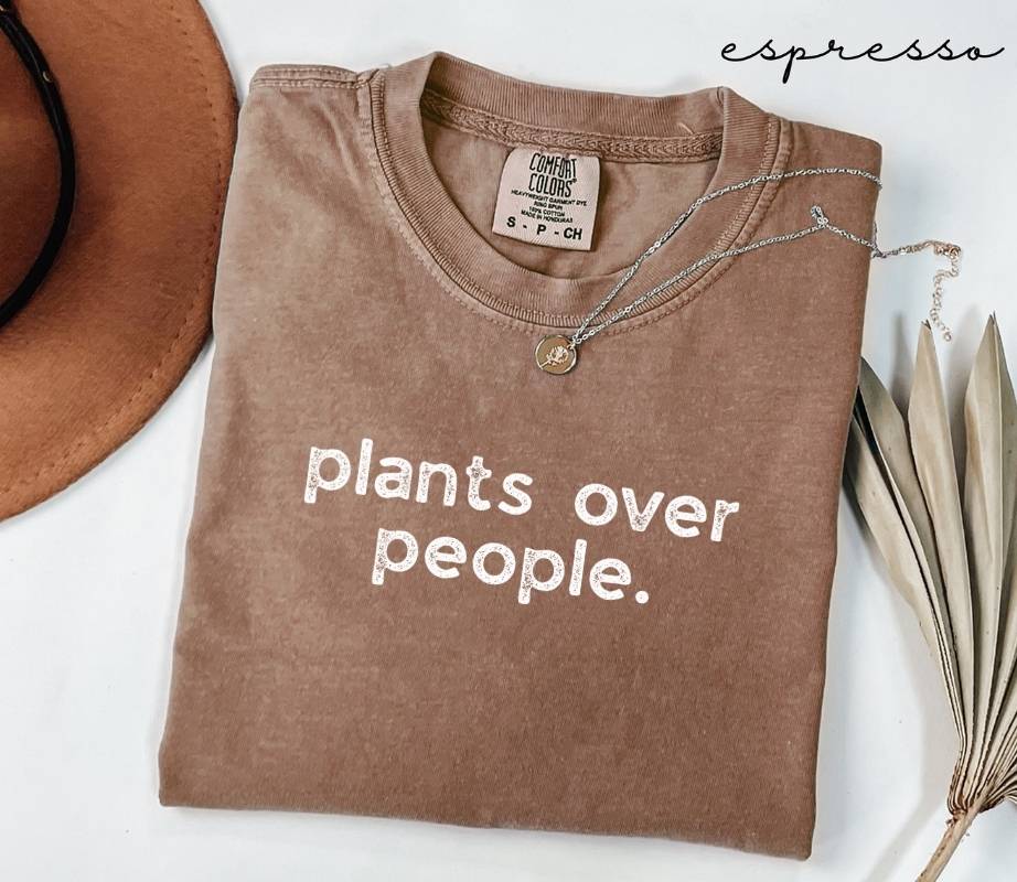 Brown (espresso) t-shirt with 'plants over people.' text, placed on a white surface with a hat and dried plant leaves.