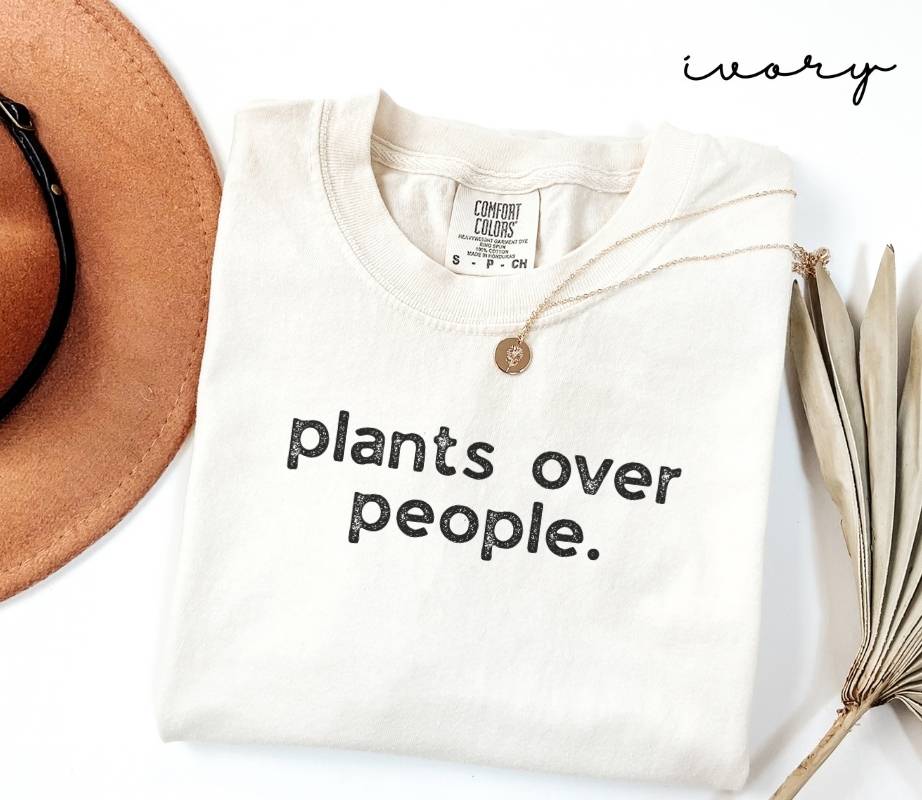 White (ivory) t-shirt with 'plants over people.' text, placed on a white surface with a hat and dried plant leaves.