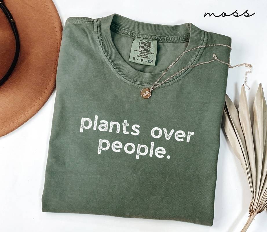 Folded green (moss) t-shirt with 'plants over people' text on a white surface with a hat and dried plant leaves.