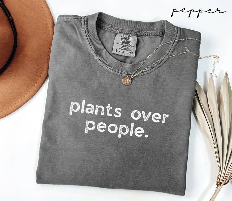 Folded gray (pepper) t-shirt with 'plants over people.' text, next to a hat and dried plant leaves.