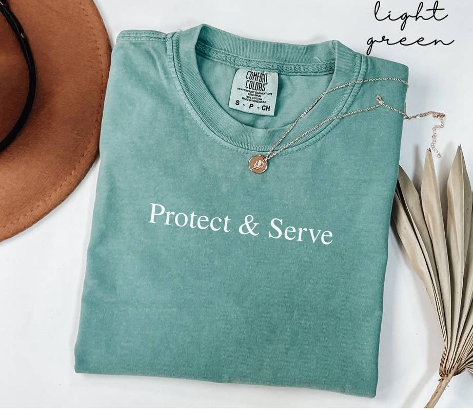 Folded light green t-shirt with 'Protect & Serve' text, worn with a necklace and hat on a white background.