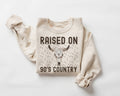 Beige (sand) sweatshirt with 'Raised on 90's Country' text and bull skull design on a white background
