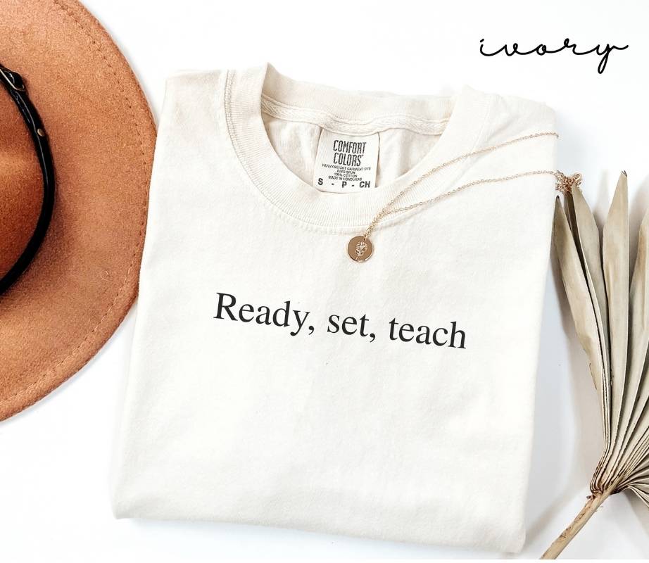 White (ivory) t-shirt with 'Ready, set, teach' text, brown hat, and dried plant on a white background