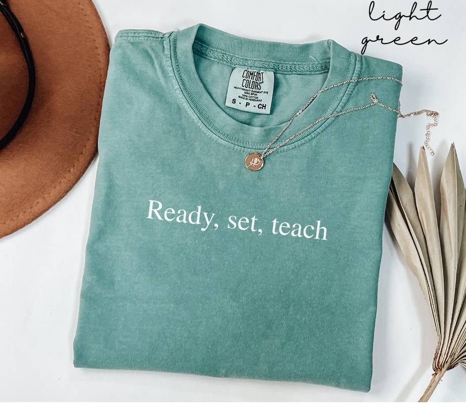 Folded light green t-shirt with 'Ready, set, teach' text on a white surface with a hat and dried leaves.