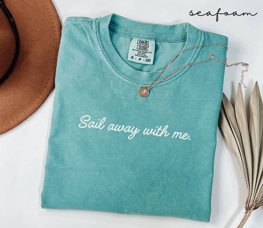 Folded sky blue (seafoam) t-shirt with the text 'Sail away with me', surrounded by a hat and dried leaves on a white background.