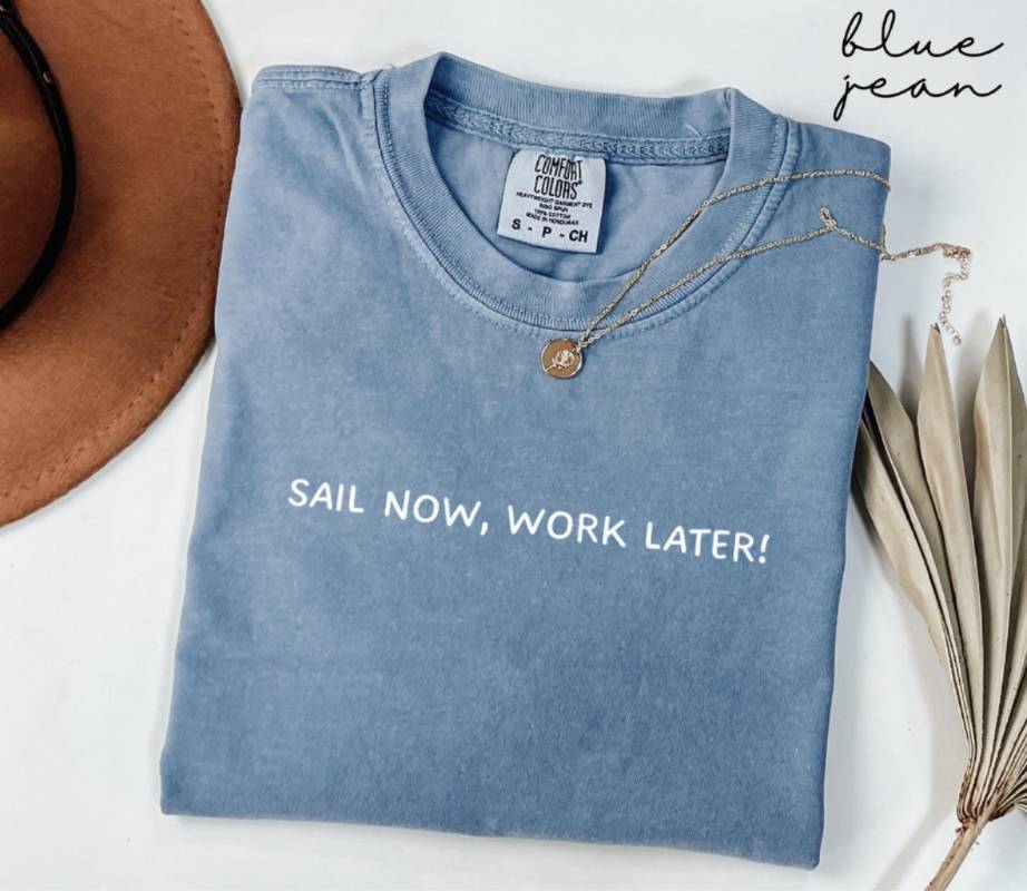 Folded blue jean t-shirt with 'Sail Now, Work Later!' text, next to a hat and dried leaves on a white background.