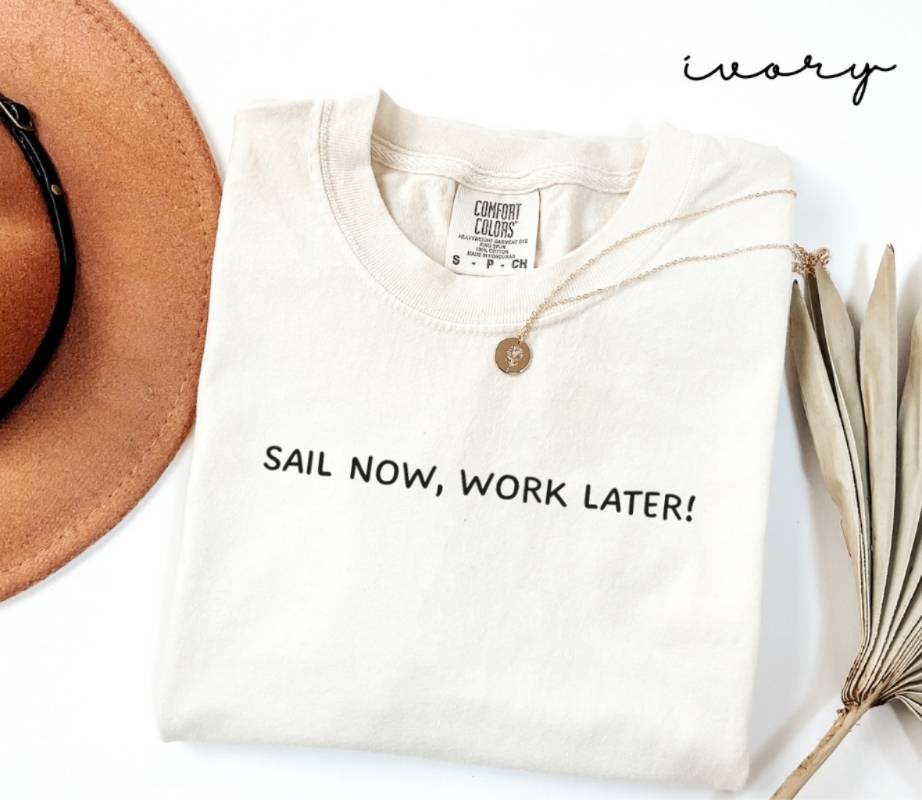 Folded white (ivory) t-shirt with 'Sail Now, Work Later!' text, next to a brown hat and dried plant on a white background.