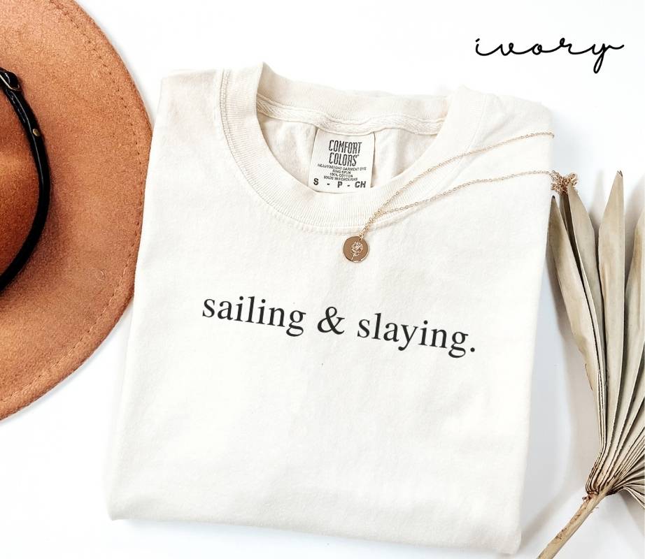 Folded white (ivory) t-shirt with 'sailing & slaying.' text, next to a brown hat and dried plant on a white background.