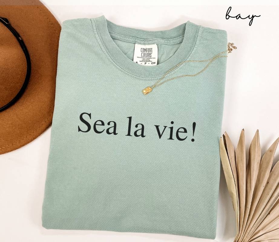Folded light green (bay) t-shirt with 'Sea la vie!' text, next to a hat and dried leaves on a white background.
