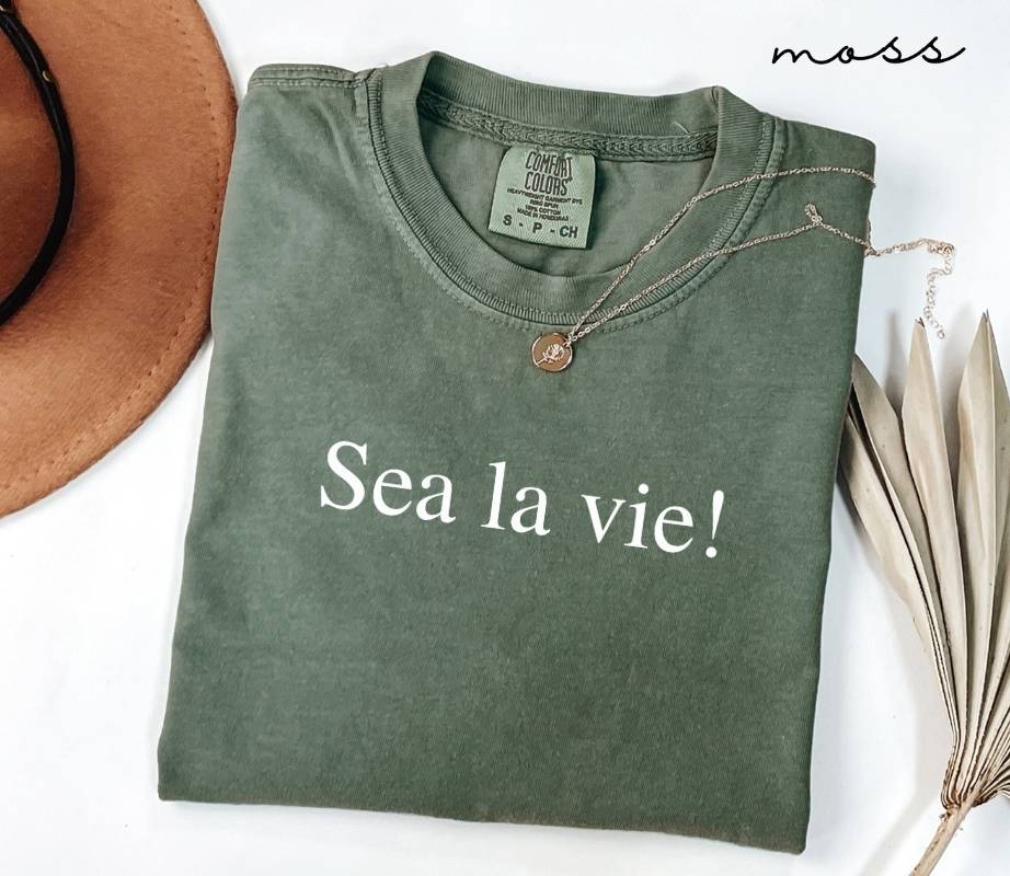Folded green (moss) t-shirt with 'Sea la vie!' text on a white surface with a hat and dried leaves.