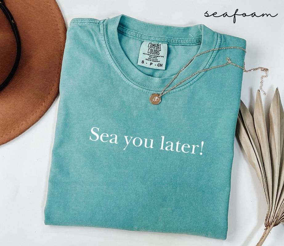 Folded teal (seafoam) t-shirt with 'Sea you later!' text, surrounded by a hat and dried leaves on a white background.