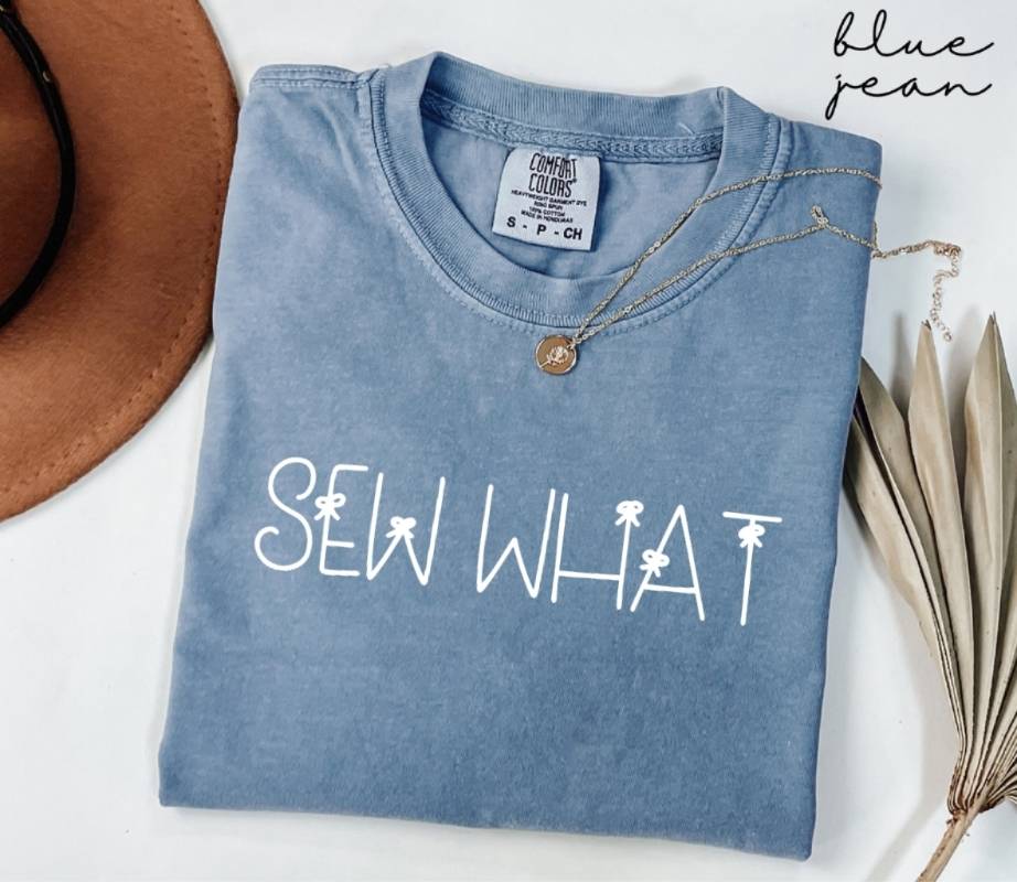 Folded blue jean t-shirt with 'SEW WHAT' text, hat, and dried leaves on a white background