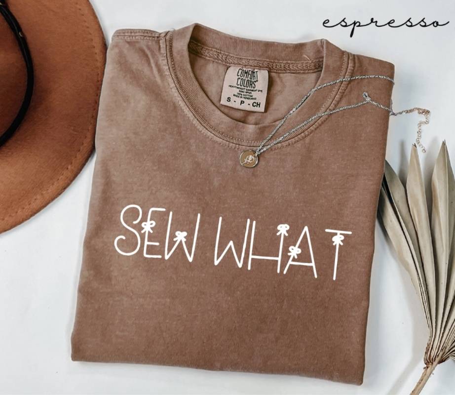 Folded brown (espresso) t-shirt with 'SEW WHAT' text, next to a hat and dried leaves on a white background.