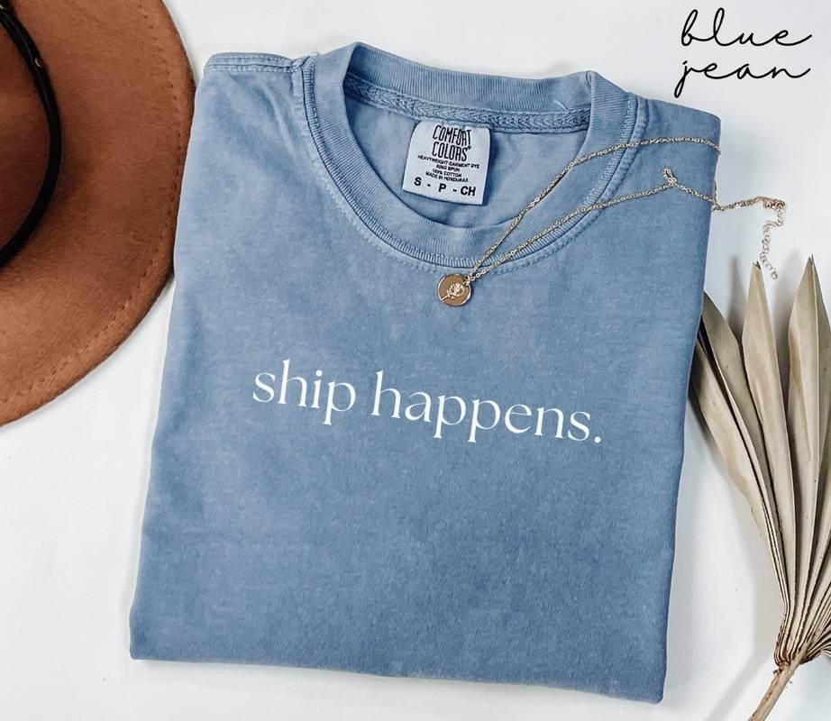 Folded blue blue jean t-shirt with 'ship happens.' text, surrounded by a hat and dried leaves on a white background.